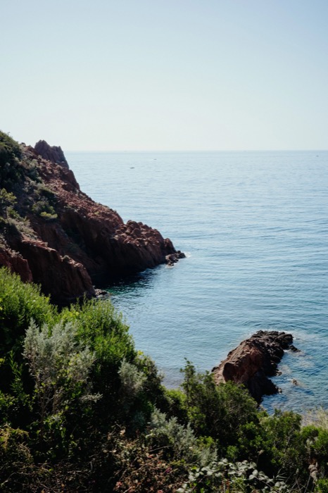 Red volcanic cliffs and scrub dropping into calm turquoise sea under a pale sky