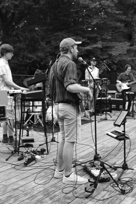 Singer in a backwards cap at a microphone on an outdoor wooden stage, band and cables around him