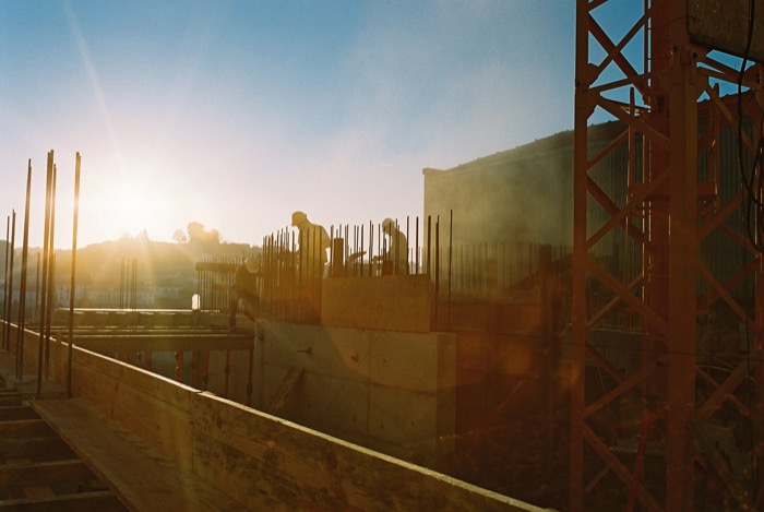 Construction workers among rebar and formwork on an unfinished concrete floor, low sun flaring across the frame