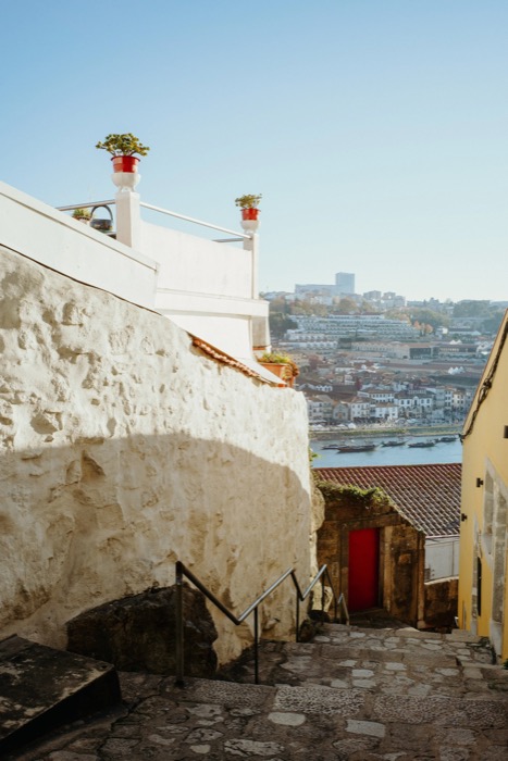 Cobbled steps beside a whitewashed wall leading down toward a red door, tiled roofs and a river town beyond