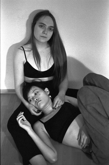 Two women in black against a bare wall, one resting her head in the other's lap