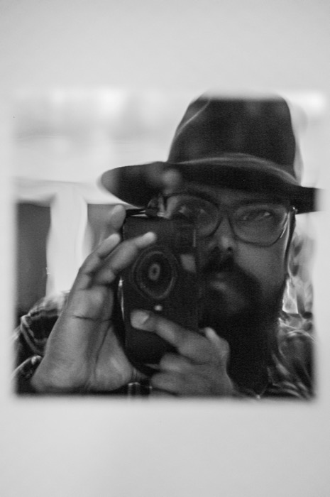 Soft-focus mirror self-portrait: man in a wide-brimmed hat and glasses raising a camera to his face