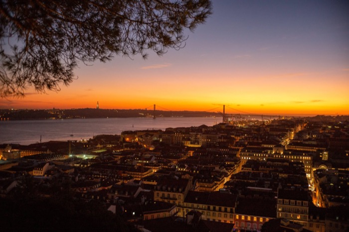 Hilltop view over a lit city at dusk, orange sky behind a suspension bridge and wide river
