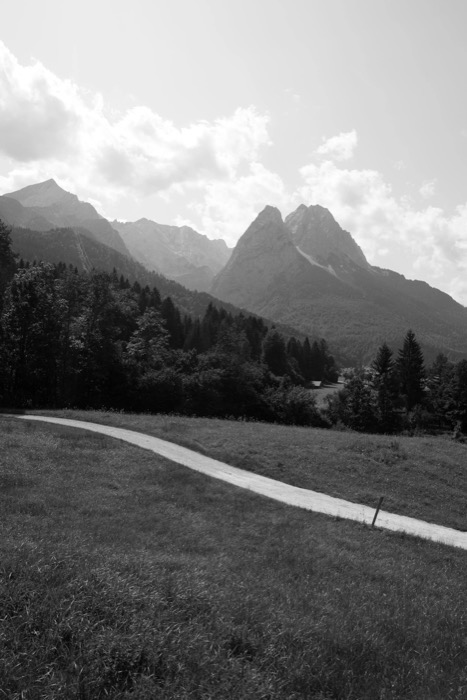 Gravel path curving through a meadow toward a twin-peaked mountain above wooded slopes
