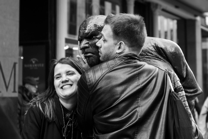 Man in a leather jacket poses cheek to cheek with a bronze living statue as a woman laughs beside them