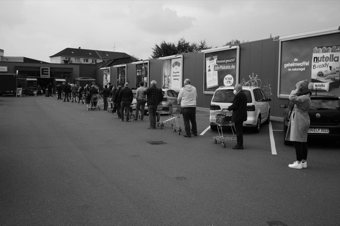 Shoppers with trolleys queuing far apart across a supermarket car park, one woman masked in the foreground