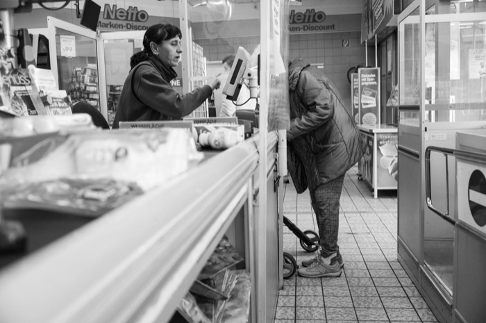 Cashier reaching over the till at a Netto discount supermarket while an elderly customer bends over her trolley