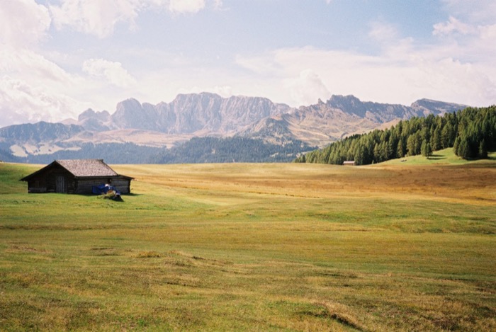 Lone wooden hay barn on a broad high alpine meadow, jagged mountain ridge and cloud beyond