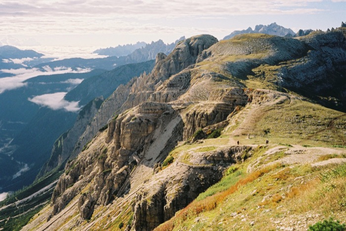 Hikers dwarfed on a ridge trail above a cloud-filled valley, jagged limestone peaks on the horizon