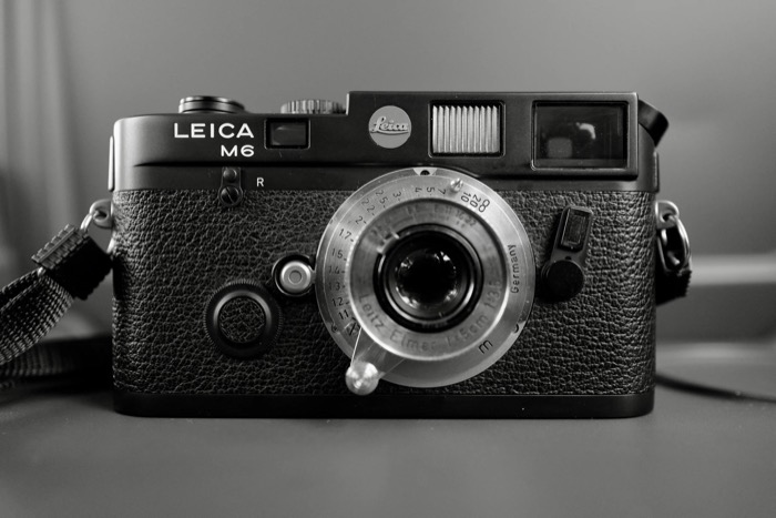 Black rangefinder camera with a small collapsible lens standing on a desk, strap coiled beside it