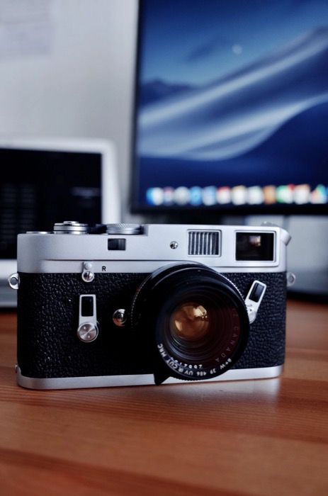 Silver rangefinder camera standing on a wooden desk in front of two glowing computer screens