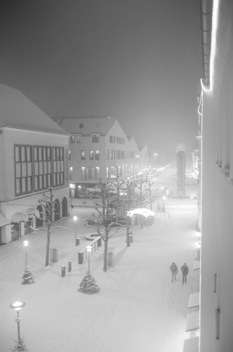Snow-covered town square at night seen from above, two figures crossing under glowing street lamps