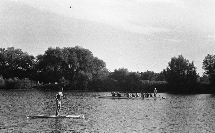 Paddleboarder glides past a rowing eight on a calm tree-lined river