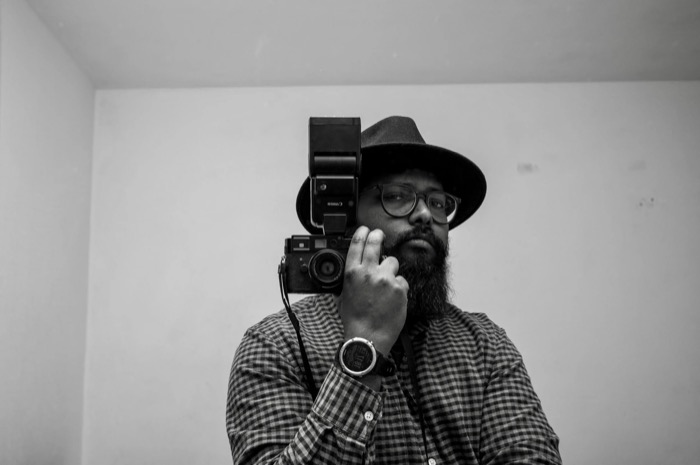 Bearded man in a fedora and checked shirt holding a flash-topped camera beside his face against a plain wall
