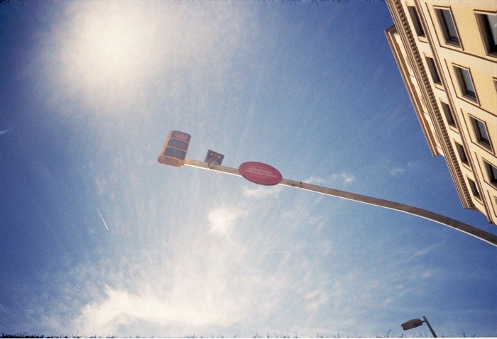 Traffic light and a red "Control Fotogràfic" sign on a tilted mast against the sun and a corner building