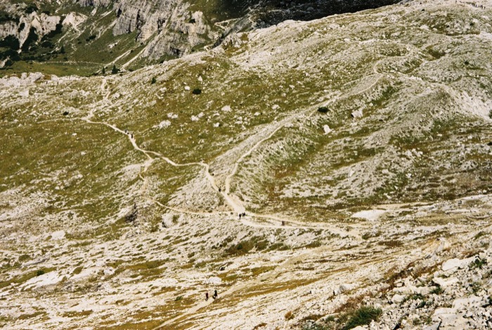 Hikers reduced to specks on braided trails crossing a vast pale scree and grass mountainside