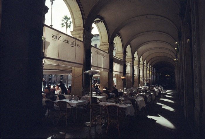 Diners under a long vaulted arcade, awnings reading les Quinze Nits, sunlight striping the floor