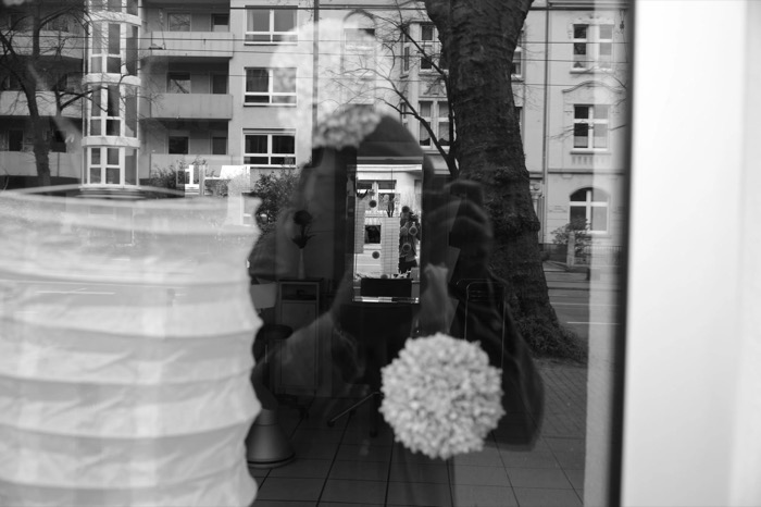 Shop window reflection layering a hooded figure, paper lanterns and the apartment blocks across the street