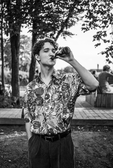 Young man in a patterned shirt drinking from a bottle under trees at an outdoor event