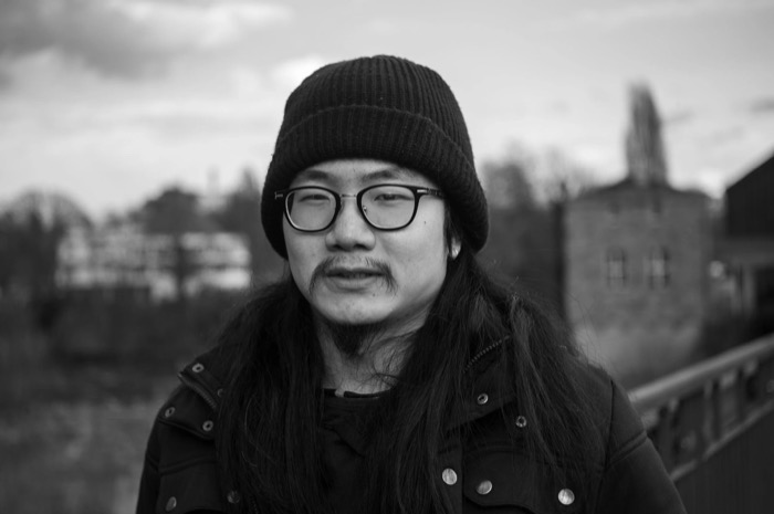 Man with long hair, moustache, glasses and black beanie half-smiling outdoors, industrial buildings blurred behind