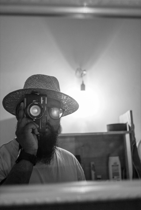 Bearded man in a straw wide-brimmed hat raising a camera to his eye, reflected in a bathroom mirror under a bare bulb